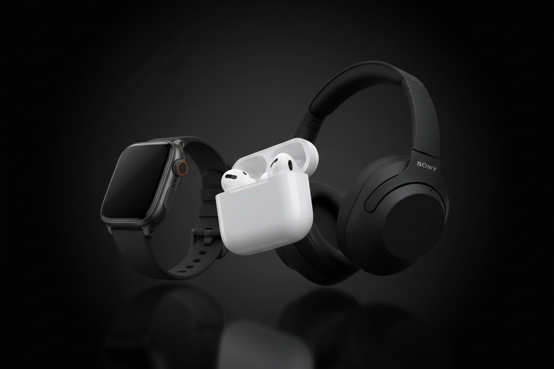 Ultra-premium product photography flat lay for an e-commerce hero banner. The image features the latest Apple AirPods Pro, a sleek black smartwatch (Apple Watch Ultra), and premium matte black wireless headphones (Sony WH-1000XM5) arranged artfully on a clean, dark gradient background. The background should be a smooth, deep black to charcoal gradient, slightly darker at the edges and brighter in the center where the products are placed. The products are slightly angled to show their design, with subtle ref